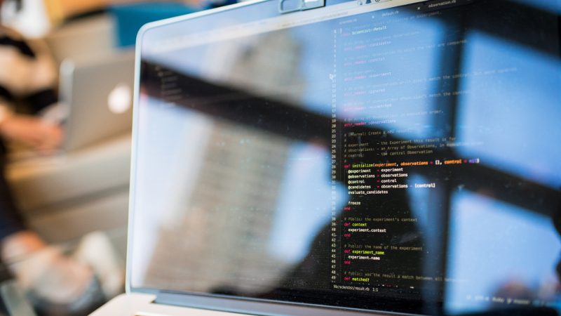 Laptop screen showing lines of programming code beside a keyboard, with a blurred background